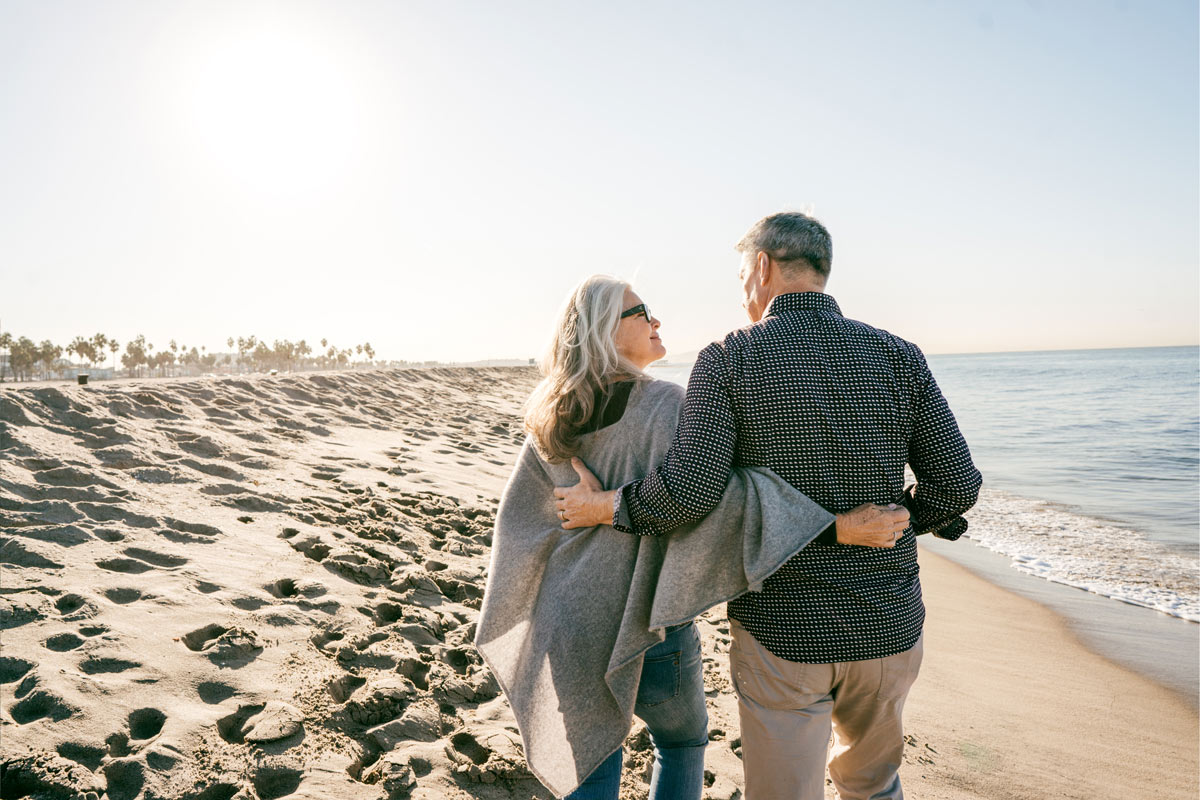 A senior couple wearing Beltone Amaze Hearing Aids walks down the beach resting their arms on each other's backs