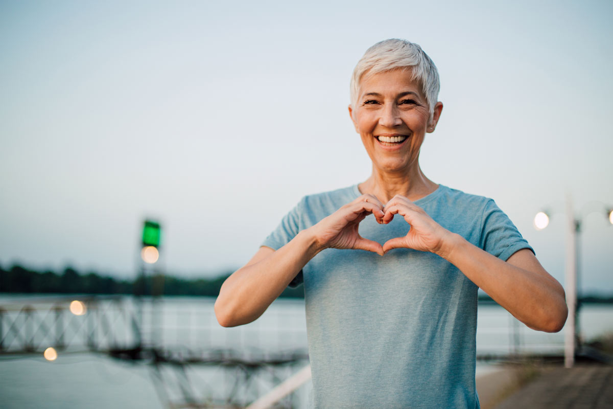 A senior woman wearing Beltone Amaze Hearing Aids stands by a river making the shape of a heart with her hands