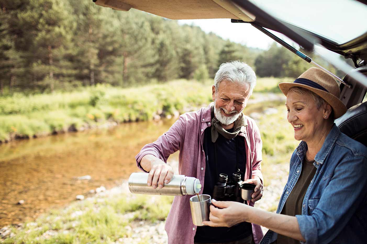 A senior couple wearing Beltone Ally hearing aids shares a cup of coffee while camping