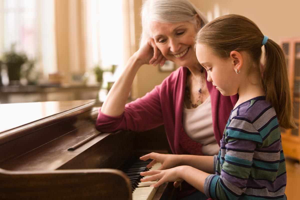 A senior woman, wearing Beltone hearing aids, sits at the piano with her granddaughter