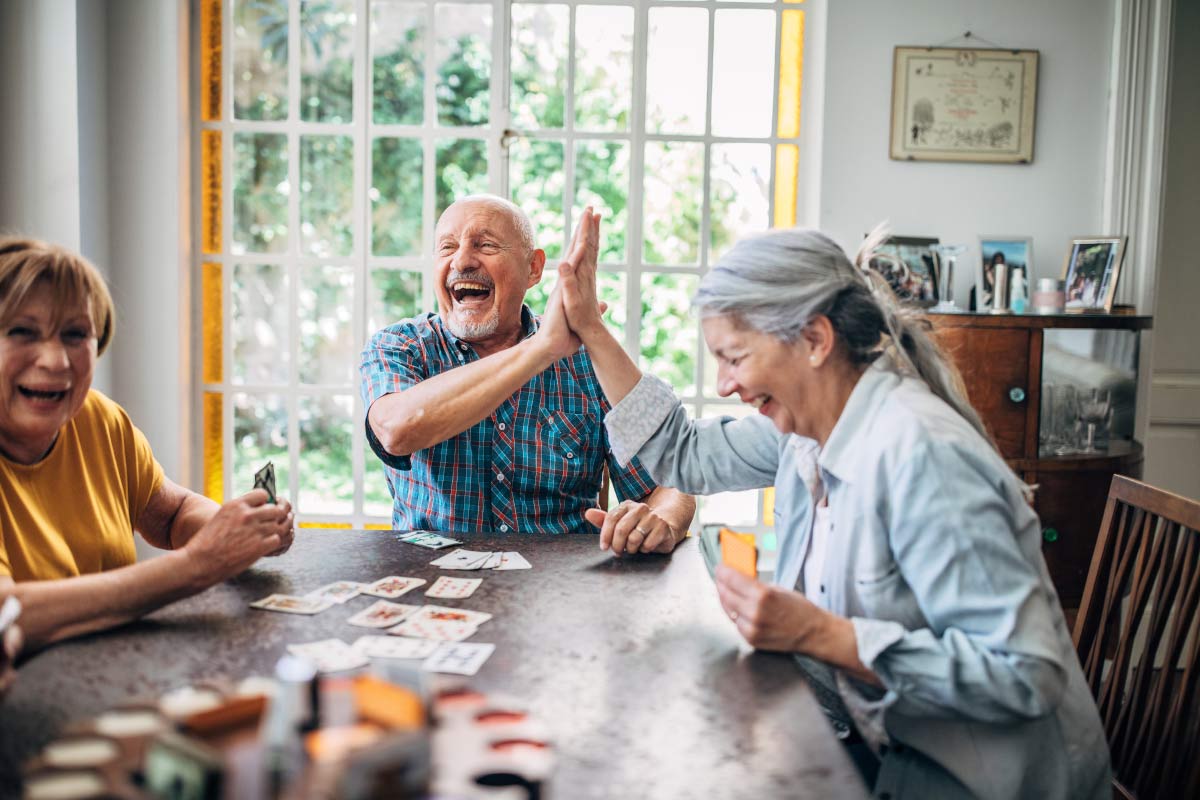 Several seniors wearing Beltone Hearing Aids play a game of cards at a kitchen table together and hi-five each other while laughing