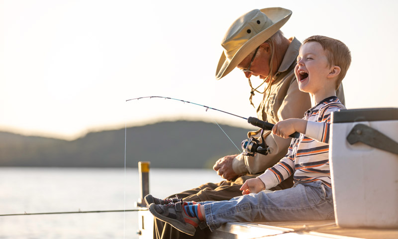 A smiling young boy sitting holding a fishing rod on a dock while his grandfather sits next to him