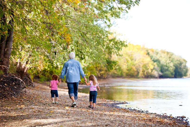 A senior walks along a river holding hands with their two grandchildren
