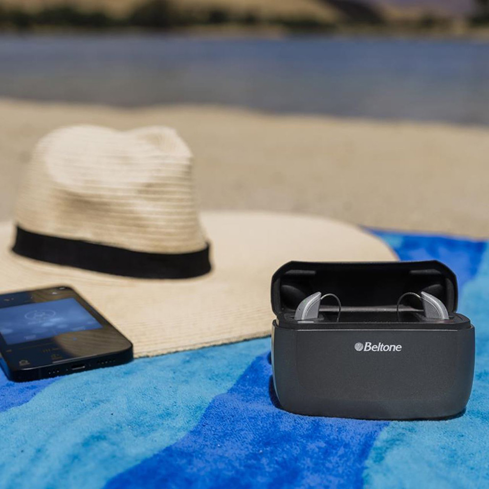 Beltone Achieve hearing aids in their recharging case next to a wide-brim hat and phone on a towel on the sand