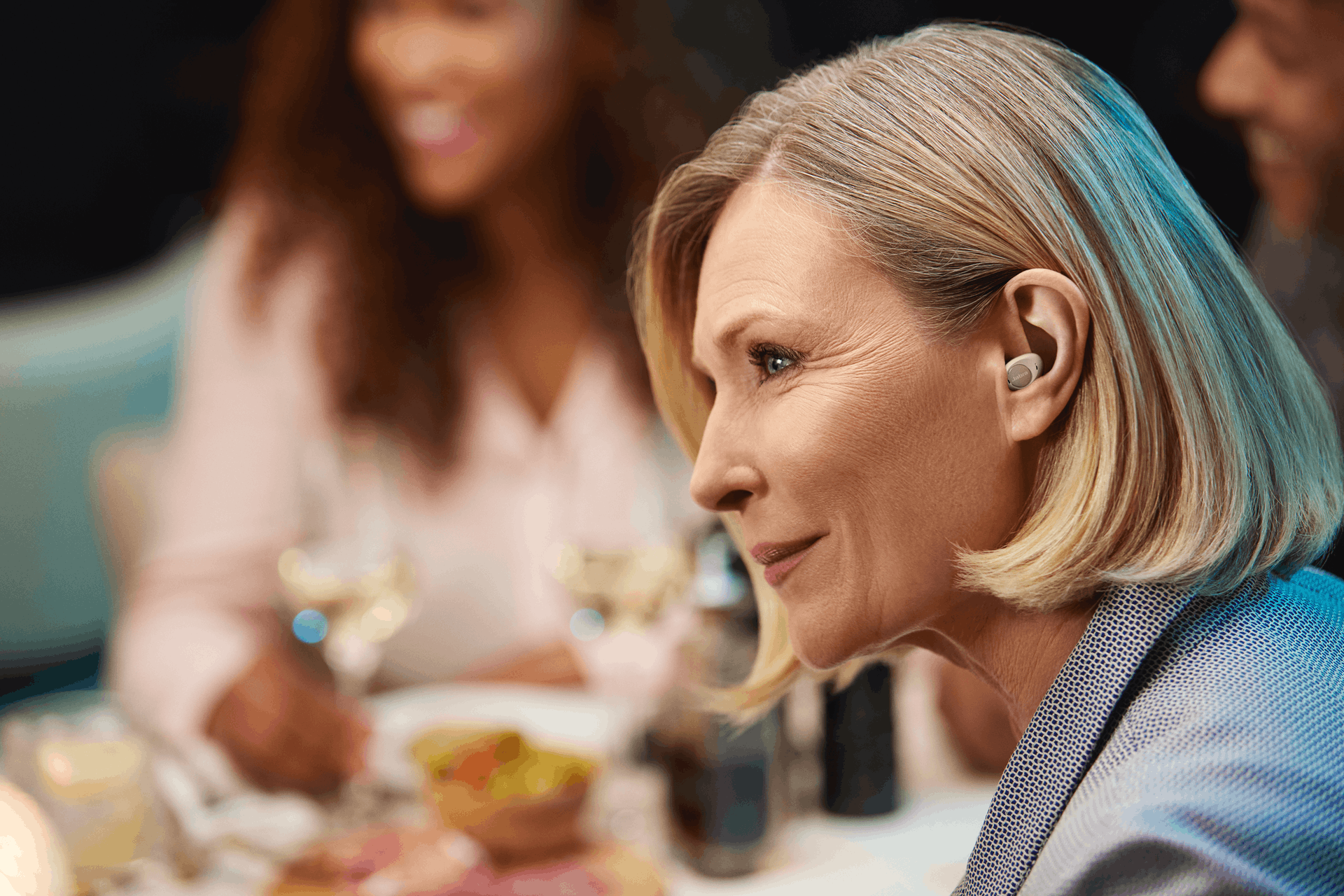 a woman wearing Jabra Enhance™ Plus earbud hearing aids listening intently to conversation around a dinner table